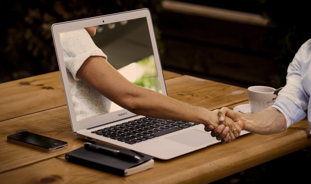 Hogyan segíti a weboldal a potenciális ügyfelek elérését és megtartását? - ehhez a blogcikkhez tartozik a kép, melyen egy kézfogás látható, az egyik kéz egy laptopból nyúlik ki